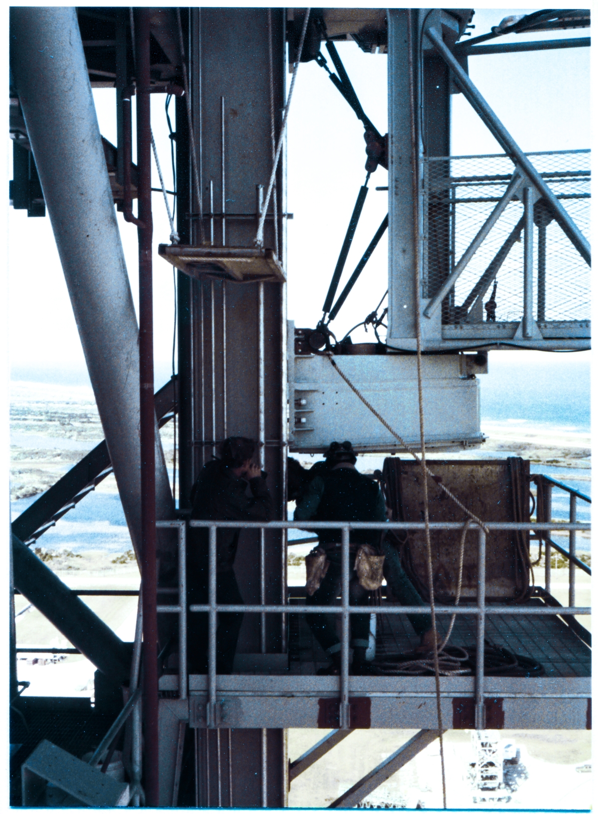 Image 115. Viewed from the GOX Arm Upper Latchback Access Platform, a very judicious choice for exact viewing angle permits us to see the GOX Arm Strongback Columns, and the GOX Arm Lower Hinge Box Connection Plates, from precisely side-on. That the Upper Latchback Access Platform existed where it did on the Fixed Service Structure, high above the Pad Deck at Space Shuttle Launch Complex 39-B, Kennedy Space Center, Florida, was most fortuitous, and our photographer did not squander this opportunity to let us see just how close we are to having the the GOX Arm finish-bolted to the tower. Union Ironworkers working for Ivey Steel Erectors, have at this point gotten the Arm into a position where the holes in the Hinge Box Connection Plates and the holes in the Strongback Columns they attach to are sufficiently lined-up to permit bolts to be inserted through them without damaging the threads, and nuts screwed on to the ends of the bolts, only finger-tight at this time, and not yet fully spun down the bolt threads and properly torqued to complete the attachment process. A knife-edge of daylight is visibly showing between the Strongback and the Hinge Box, and interrupting that knife-edge of daylight, the shanks of the bolts are clearly visible, in-place. The heads of the bolts, on the left side, are obscured from view by a run of conduit, which is attached to the back side of the Strongback Column Flange, but on the other end of the bolts, we can clearly see the nuts in place, as well as the washers which will rest beneath them once they are finish-torqued. Elsewhere on the Upper and Lower Hinge Boxes, out of frame and unseeable in this photograph, work continues with the peculiar combination of overwhelming brute force and a surgeon's delicate finesse which you only get with ironworking, causing the rest of the bolt holes to come into proper alignment, which will then allow for the GOX Arm to at-last get securely and permanently fastened to the FSS, where it can do its job supporting Space Shuttle launches. Photo by James MacLaren.